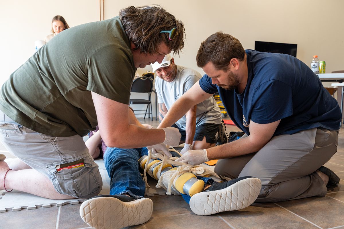First-aid trainees in a classroom practice bandaging an injured person's legs while two responders kneel beside them, creating a focused mood.