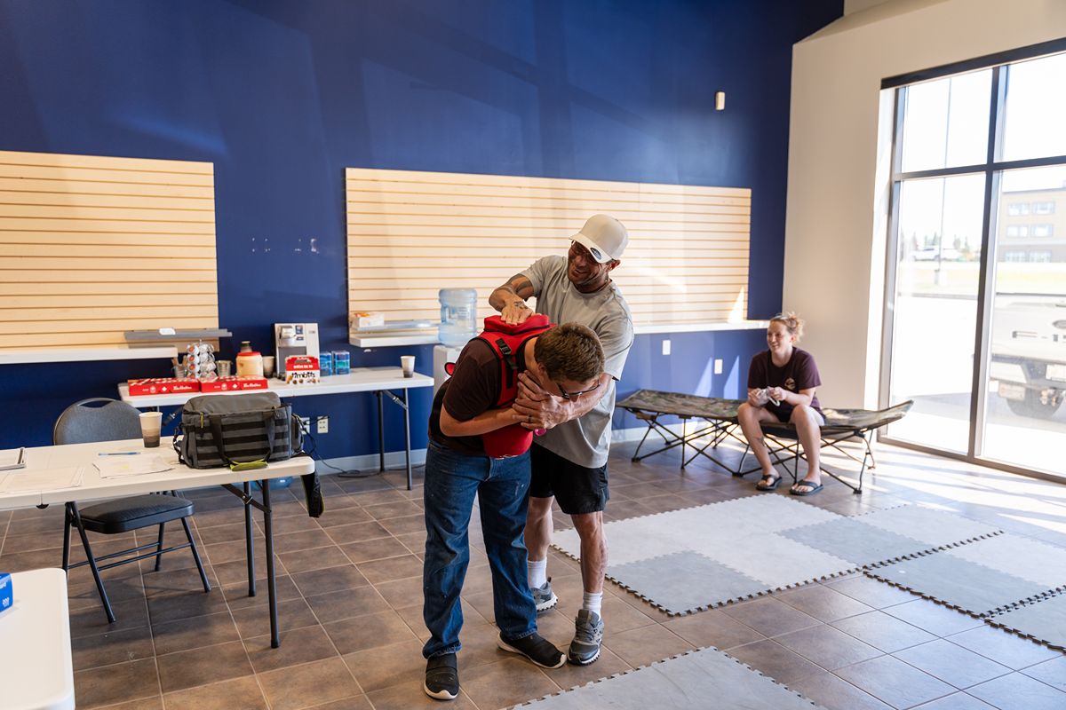 In a bright training room, an instructor supports a bent participant during a first-aid exercise while another person observes nearby.