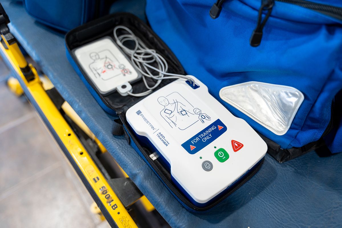 Portable automated external defibrillator (AED) in an open case on a blue medical stretcher, beside emergency equipment; clinical, ready tone.